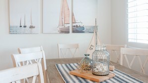 Comedor con mesa de madera, sillas blancas y un mantel a rayas. Hay tres cuadros en la pared que muestran veleros. En la mesa, una botella de vidrio con tapón de metal y objetos decorativos.