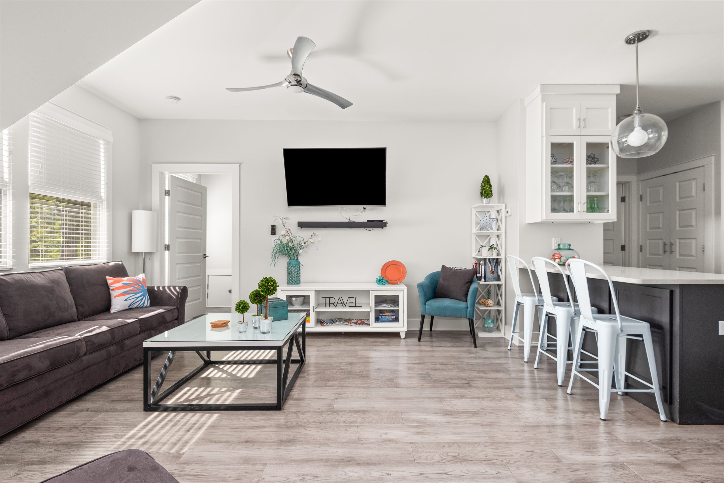 A modern living room with a sofa, coffee table, and TV. There is a dining area with chairs and a kitchen with cabinets and a fridge.