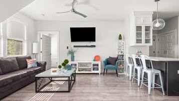 A modern living room with a sofa, coffee table, and TV. There is a dining area with chairs and a kitchen with cabinets and a fridge.