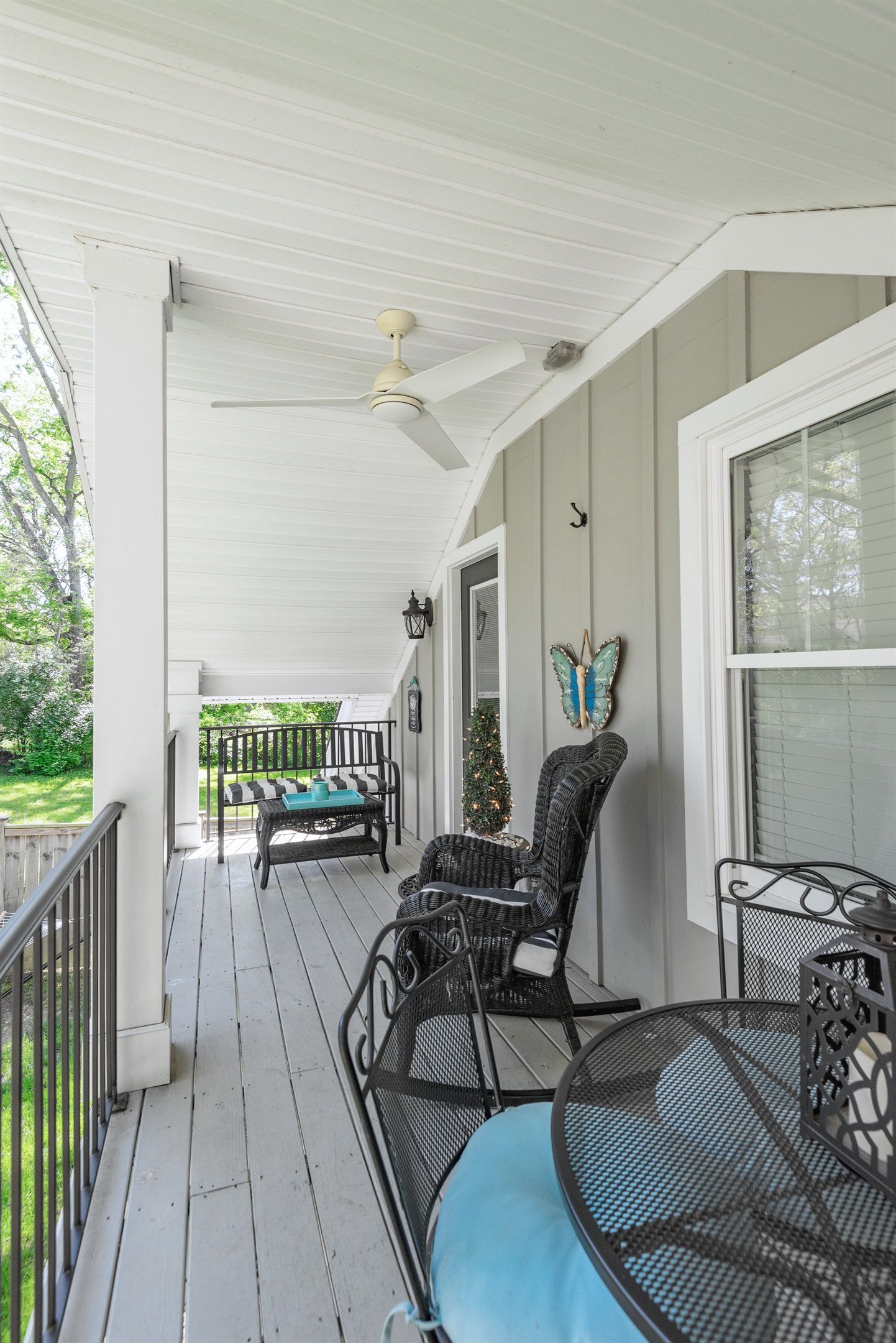 A covered porch with a ceiling fan, a black metal table, and two wicker chairs with cushions.