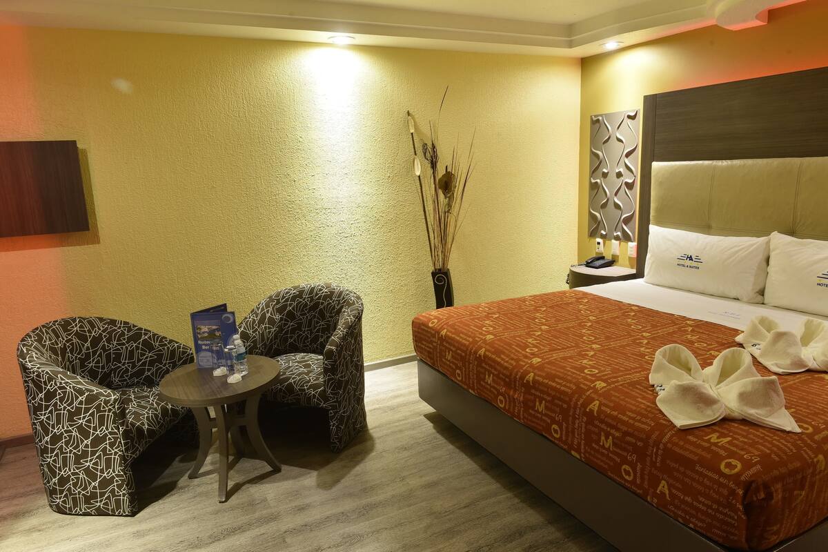 A hotel room with a bed, two patterned armchairs, a small table with a brochure, and a vase with dried plants.