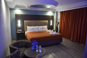 Quarto de hotel com uma cama grande, uma mesa redonda com um cardápio e uma mesa de cabeceira com abajur.