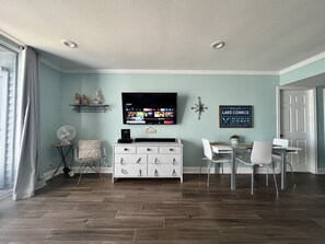 A modern living room with a flat-screen TV, a white entertainment unit, and a dining area with white chairs and a glass table.