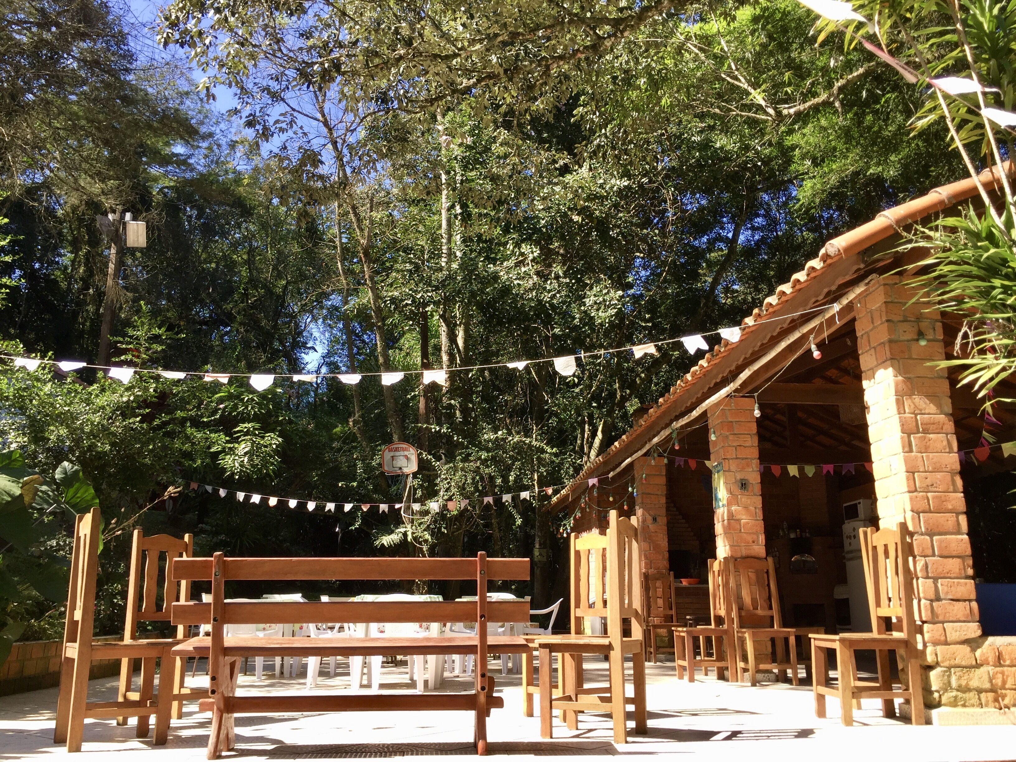 An outdoor seating area with wooden benches and tables, surrounded by trees and a brick building.