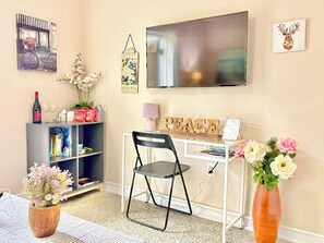 Ein Zimmer mit weißem Schreibtisch, schwarzem Stuhl, Bücherregal, Wand-Fernseher und einer Blumenvase.