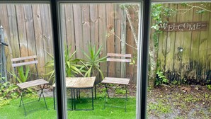 On aperçoit, à travers une fenêtre, un jardin avec une clôture en bois, un panneau d’accueil, ainsi que deux chaises et une table en bois.