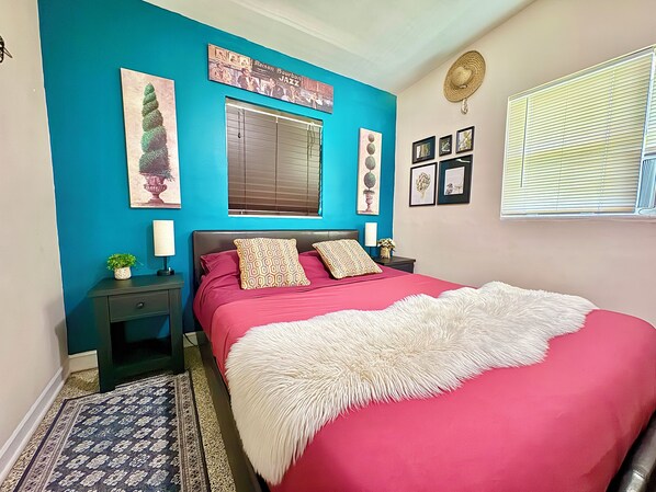 A bedroom with a bed, a nightstand, a window with blinds, a wall clock, and framed pictures.