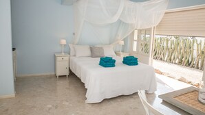 A bedroom with a bed, nightstands, a window with a view of cacti, and a table with a glass container. - Hakuna Matata Bonaire (Kralendijk)