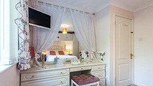Ein Schlafzimmer mit weißem Schminktisch, einem Bett mit weißer Kopfteil sowie einem Fenster mit floralem Vorhang und einer geschlossenen Tür.