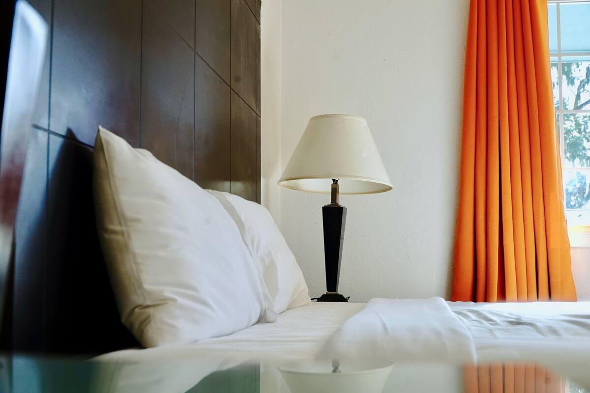 A hotel room with a bed, a lamp, a window with curtains, and a view of trees.