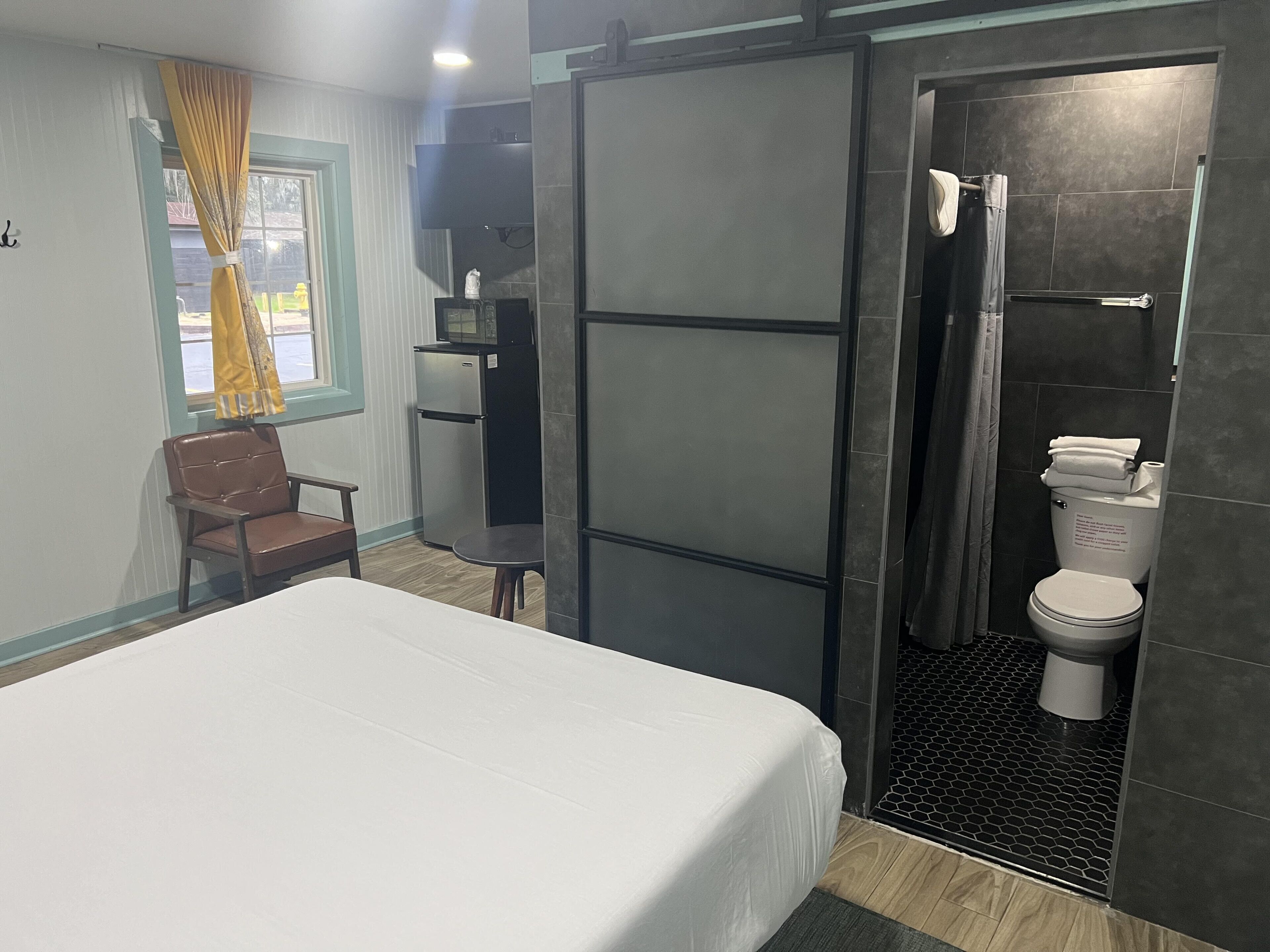 A bedroom with a bed, a chair, a small table, a refrigerator, and a bathroom with a toilet and shower.