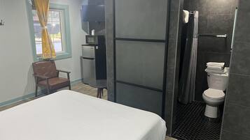 Un dormitorio con cama, una silla, una mesita, refrigerador y baño completo con inodoro y ducha.