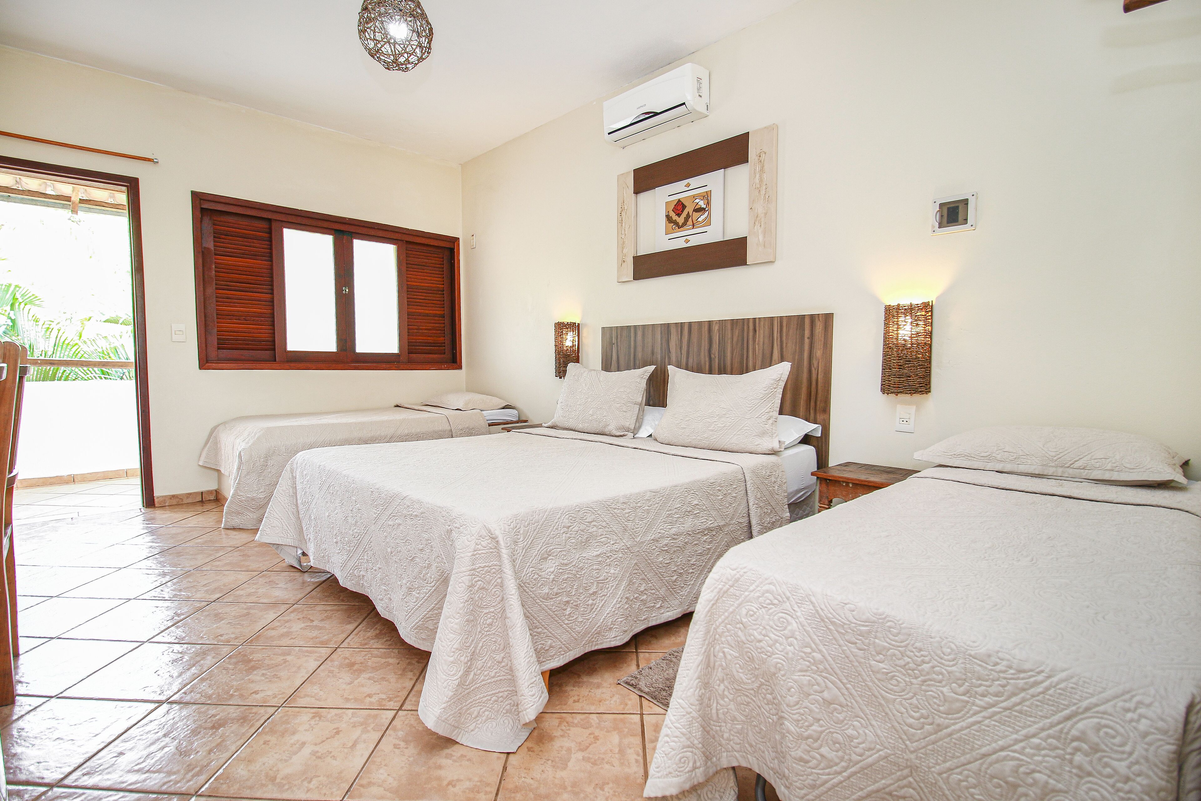 Quarto de hotel com três camas, uma janela grande com persianas de madeira, um aparelho de ar condicionado e uma luminária fixada na parede.