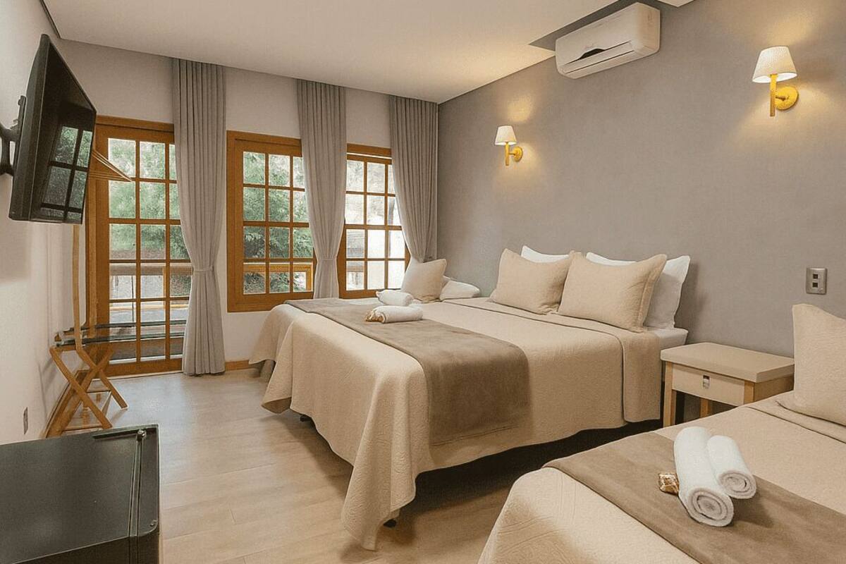 A hotel room with two beds, a TV, an air conditioning unit, and a window with curtains.