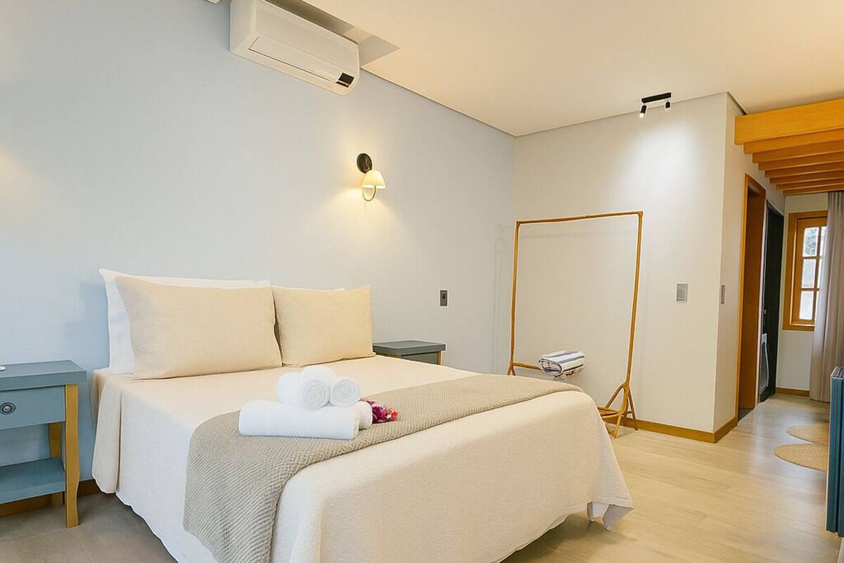 A modern hotel room with a neatly made bed, a bedside table, a wall-mounted air conditioner, and a standing mirror.