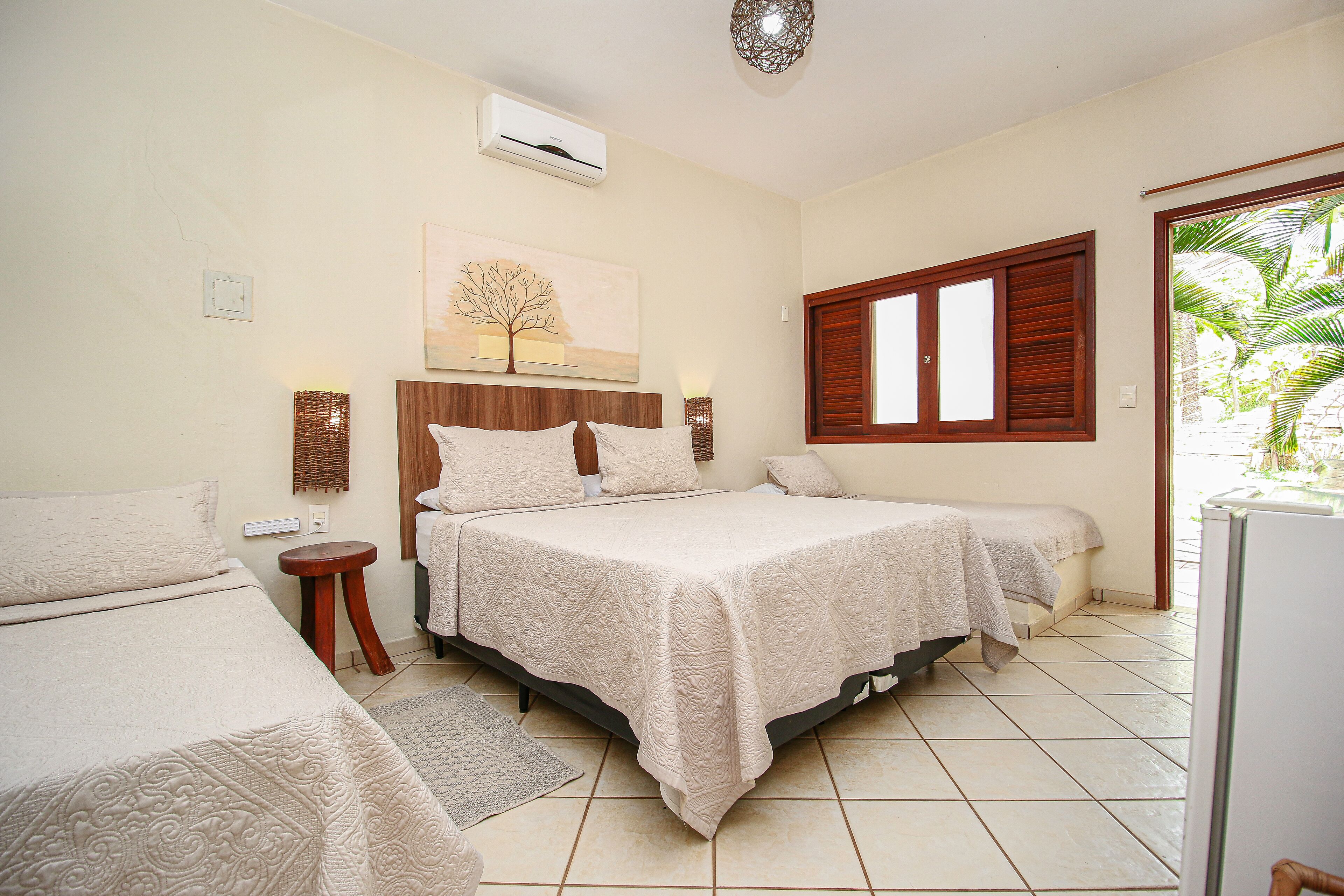 A bedroom with two beds, a wooden headboard, a wall-mounted air conditioner, a window with wooden shutters, and a view of greenery outside.