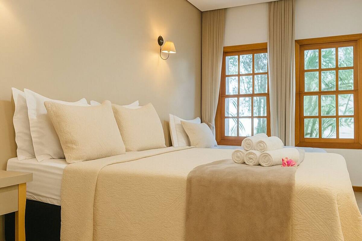 A neatly made bed with white linens and a beige throw blanket, a bedside table with a lamp, and a window with curtains.
