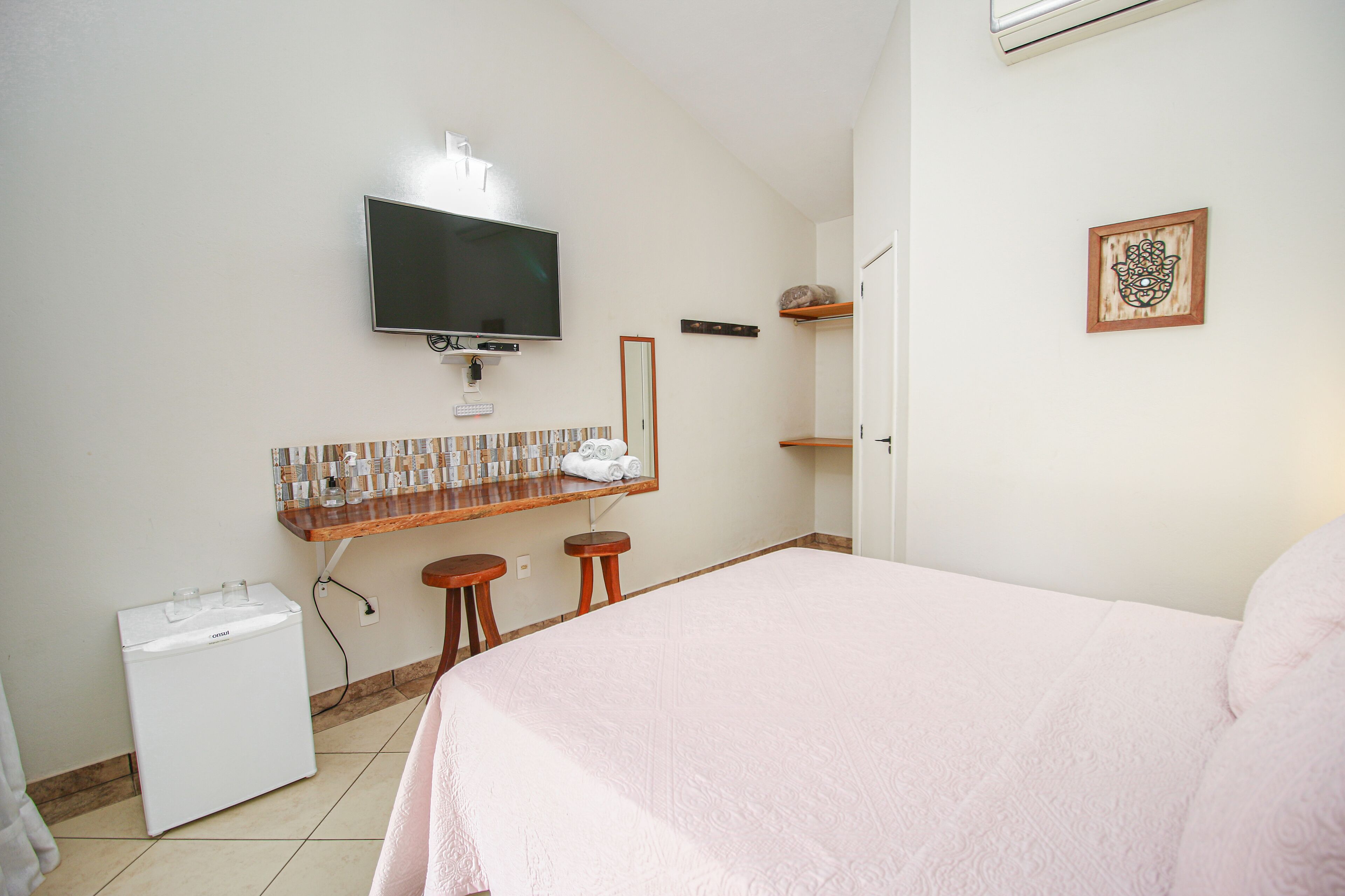 A hotel room with a bed, a TV mounted on the wall, a small refrigerator, and a wooden shelf with towels.