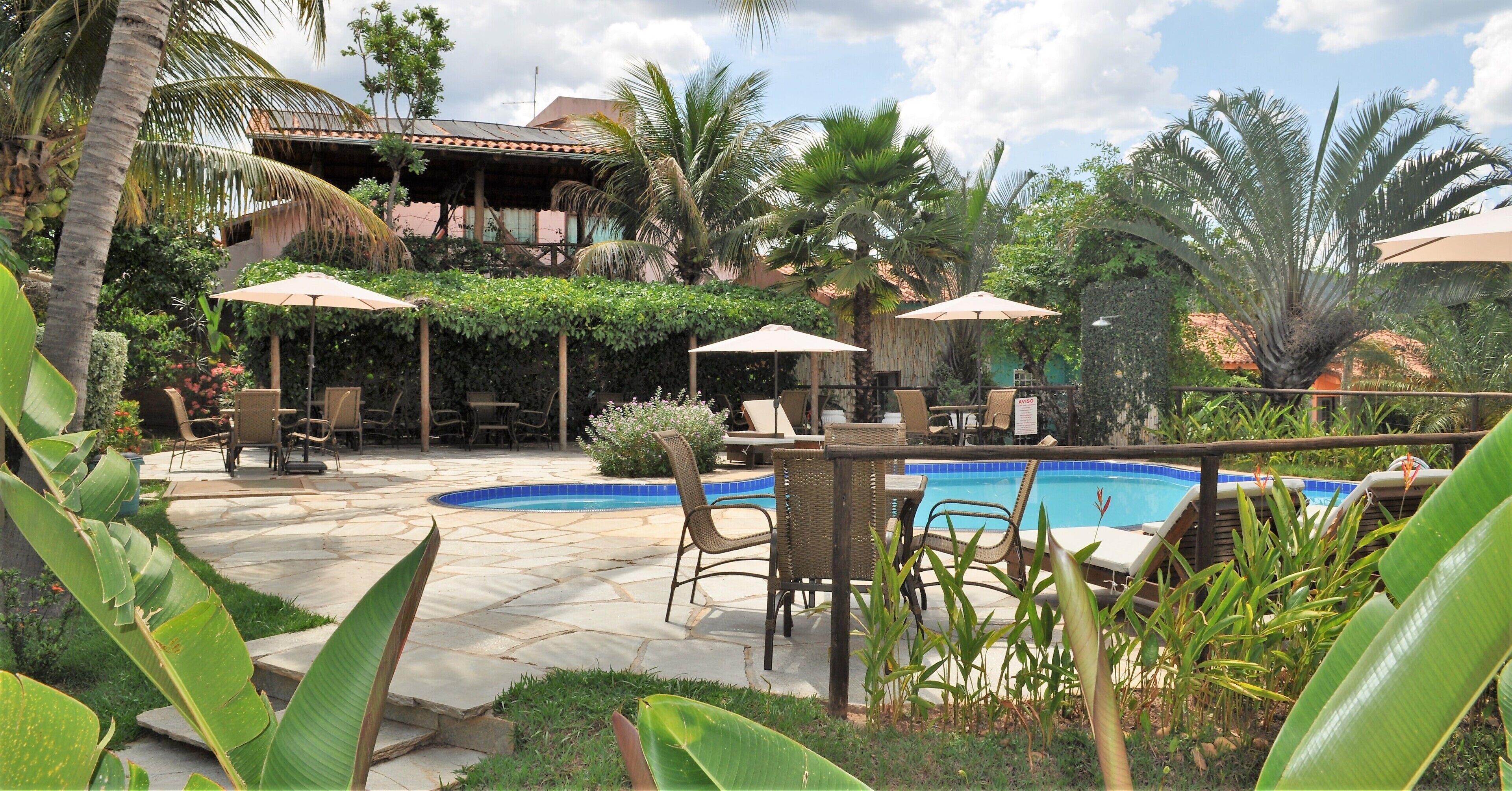 A pool area with lounge chairs and umbrellas, surrounded by tropical plants and a building with a tiled roof.