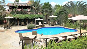 Una piscina con tumbonas y sombrillas, rodeada de vegetación y un edificio con tejado de azulejos.