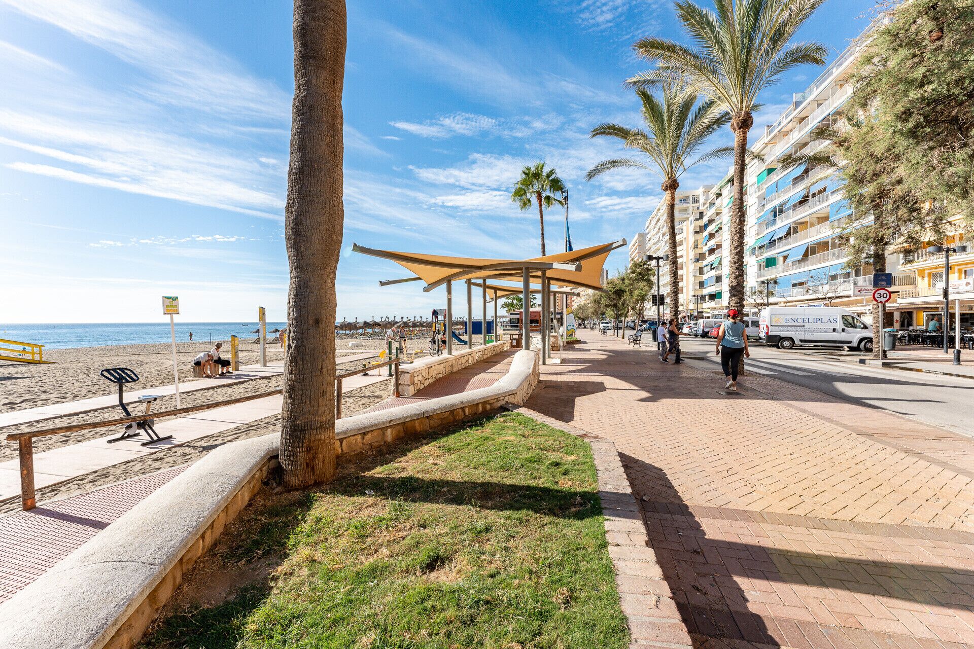 Un paseo marítimo con palmeras, una playa y una zona cubierta con asientos.
