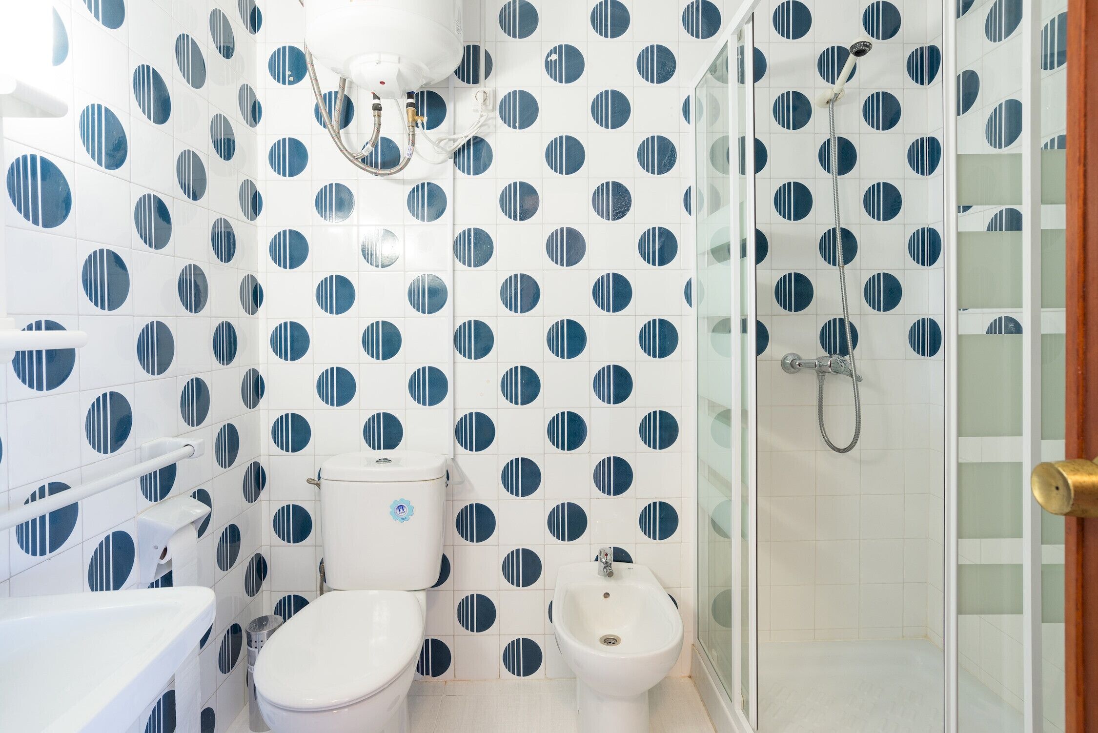 Une salle de bain avec des carreaux blancs et des motifs circulaires bleus, des toilettes, un bidet et une douche séparée par une paroi en verre.