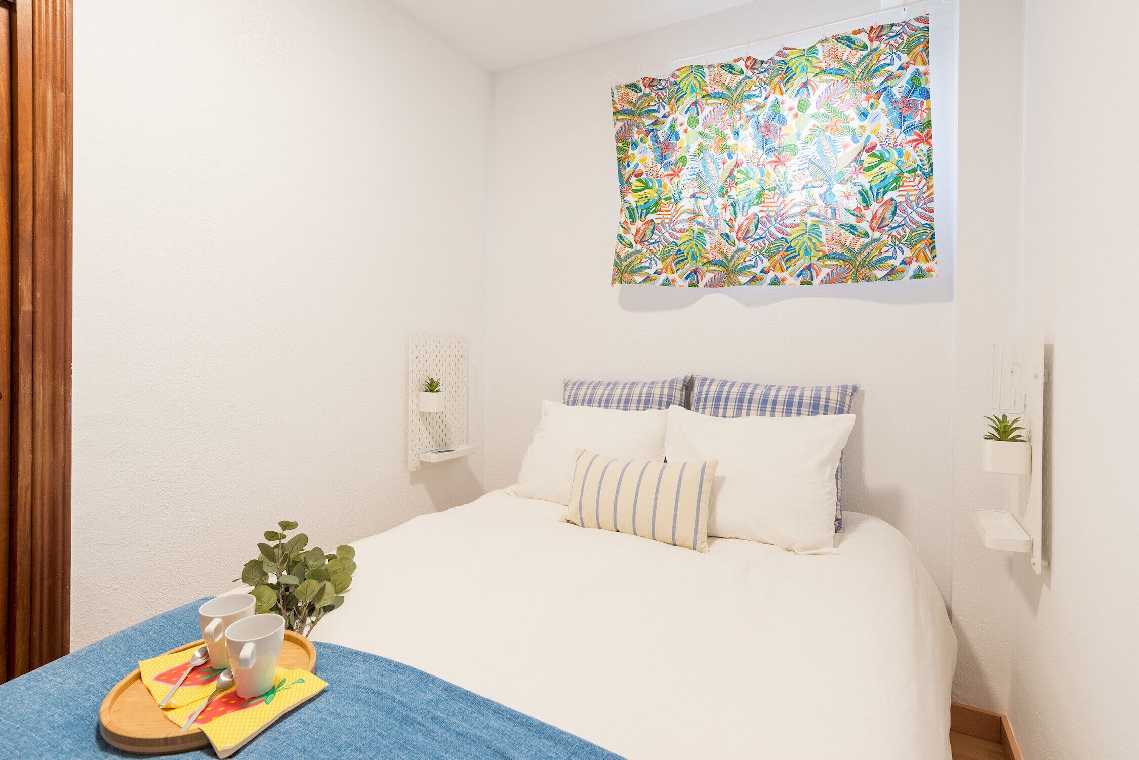 A neatly made bed with white bedding and blue and white pillows, a colorful tapestry on the wall, a small plant on a shelf, and a tray with cups and a spoon on the floor.