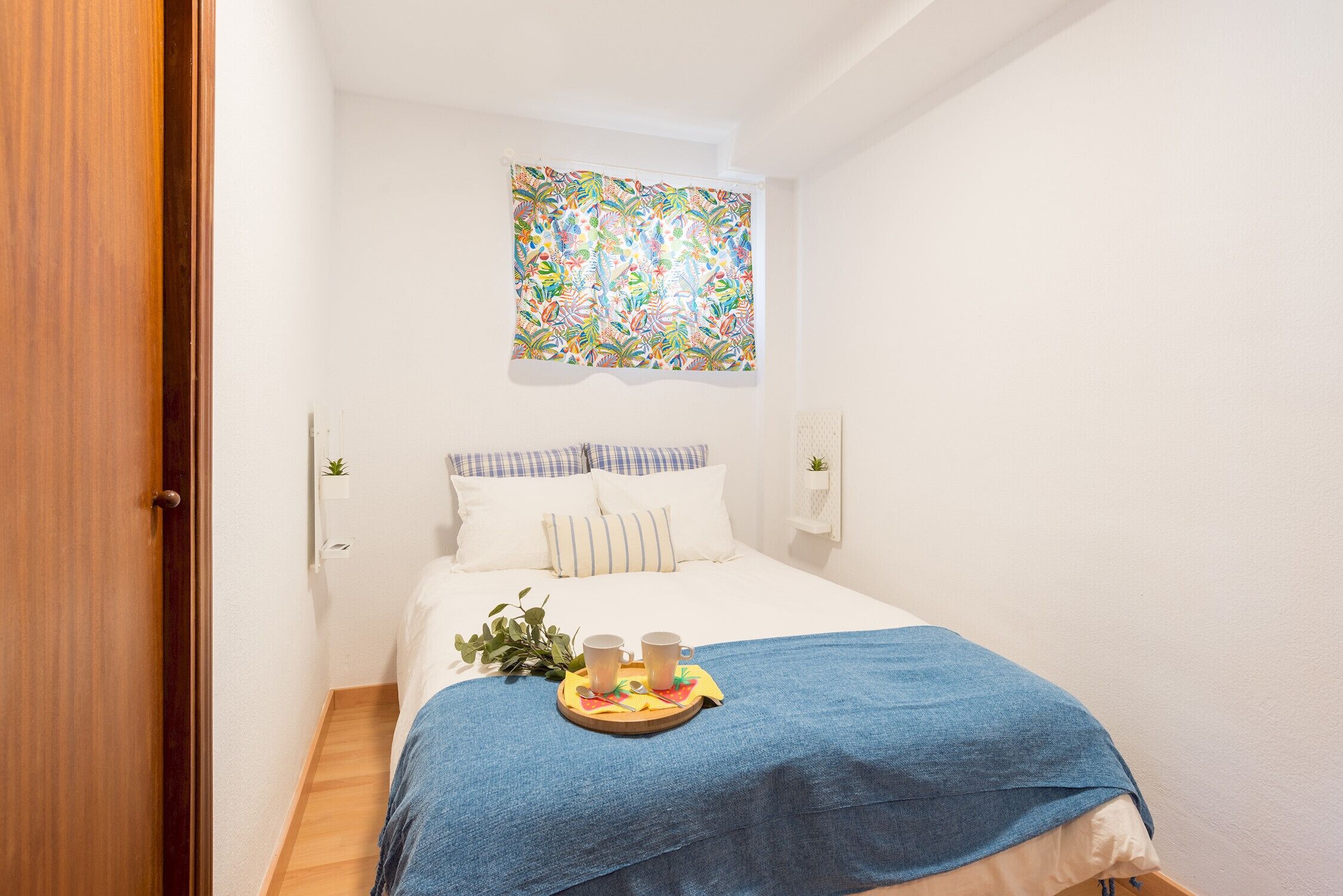 Een slaapkamer met een bed, een blauwe deken, een dienblad met twee bekers en een kleurrijke wandtapijt.