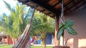 A wooden table with a woven plant pot, surrounded by wooden chairs, under a covered patio area with a hammock and palm trees in the background. - Pousada das Papoulas (Búzios)