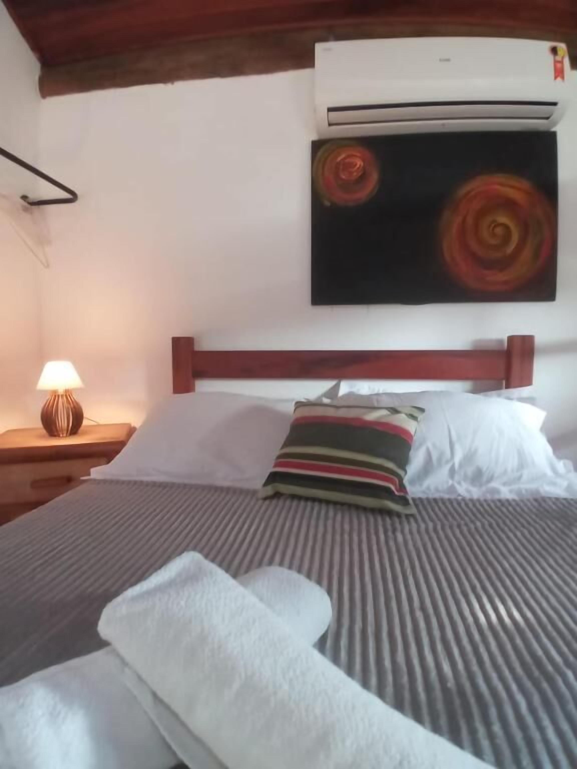 Quarto de hotel com cama, mesa de cabeceira com abajur, ar-condicionado fixado na parede e um quadro.