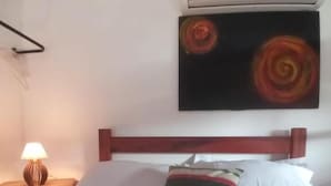 A hotel room with a bed, a nightstand with a lamp, a wall-mounted air conditioner, and a painting. - Pousada das Papoulas (Búzios)