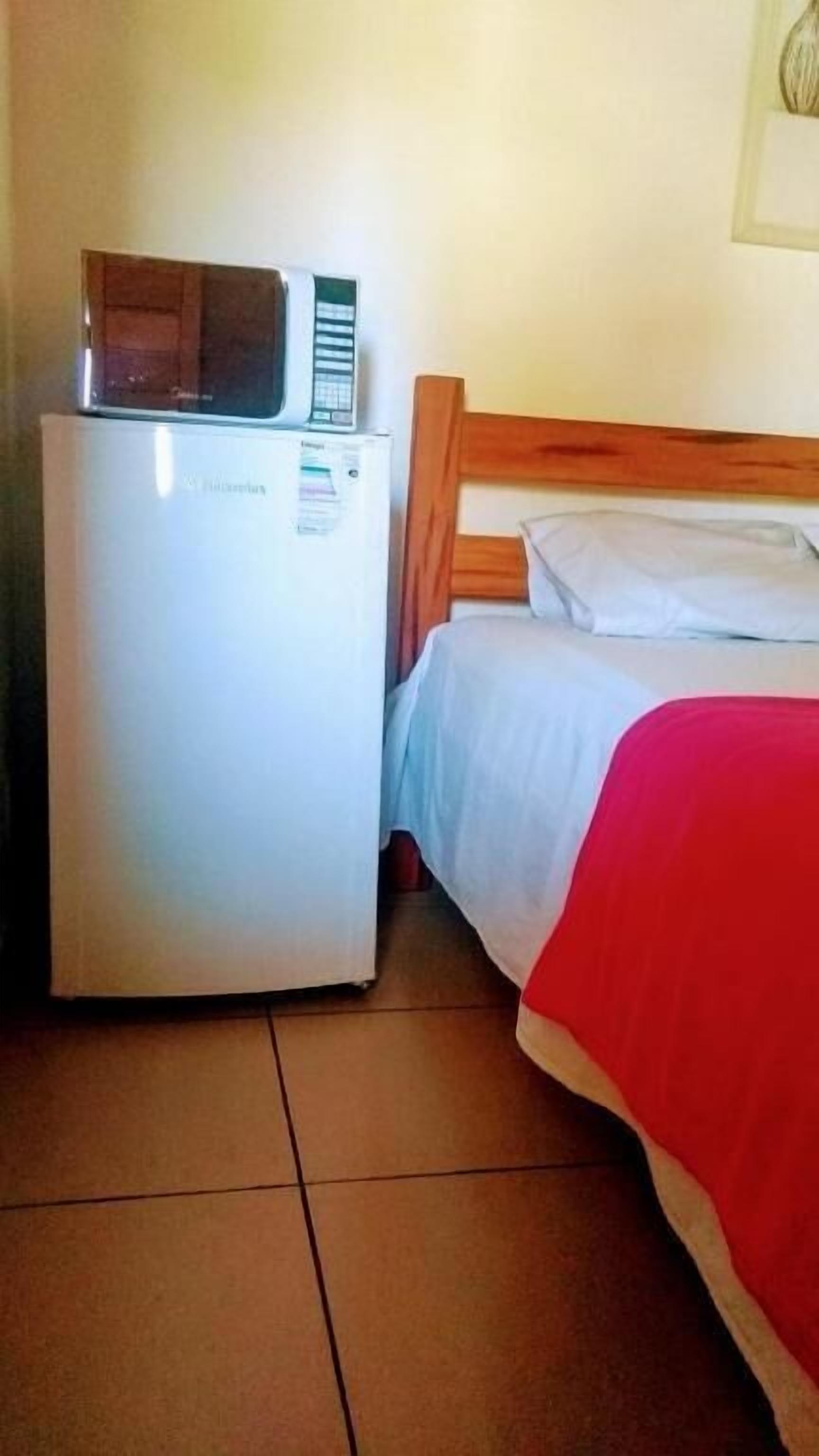 A compact room with a bed, a wooden headboard, a white wall, and a small white refrigerator with a microwave on top.