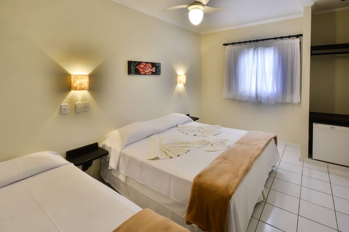 A hotel room with two beds, a ceiling fan, wall-mounted lights, a small table, a window with curtains, and a mini fridge.