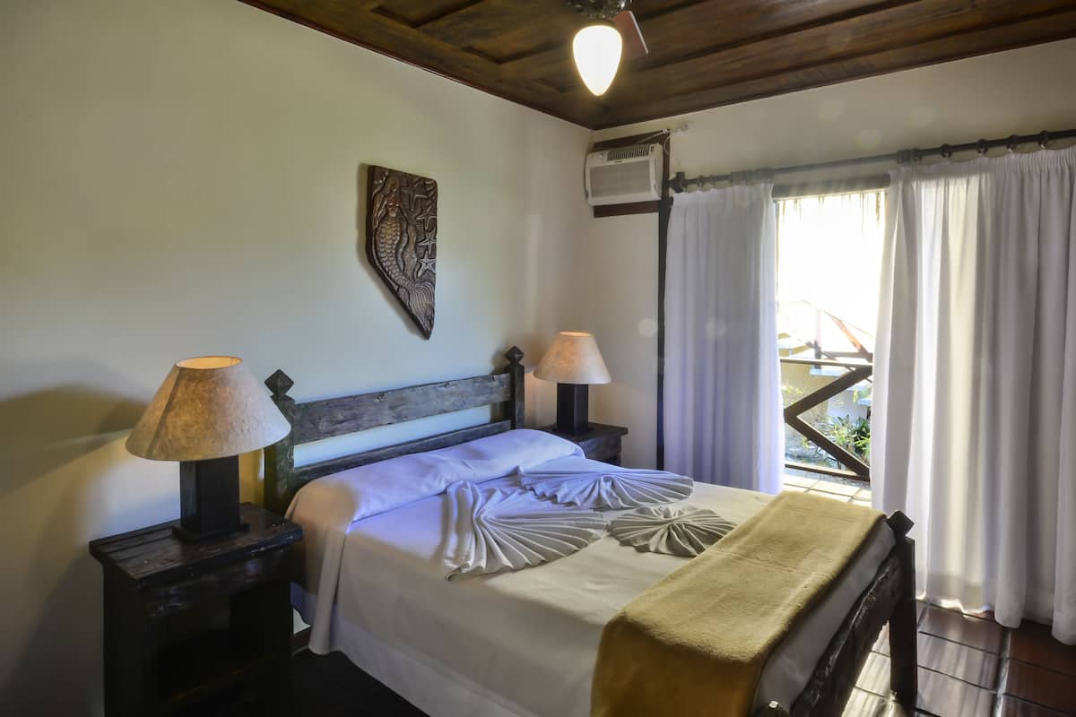 A neatly made bed with white linens and decorative pillows, two bedside lamps, a wooden headboard, and a view of the outdoors through a window with curtains.