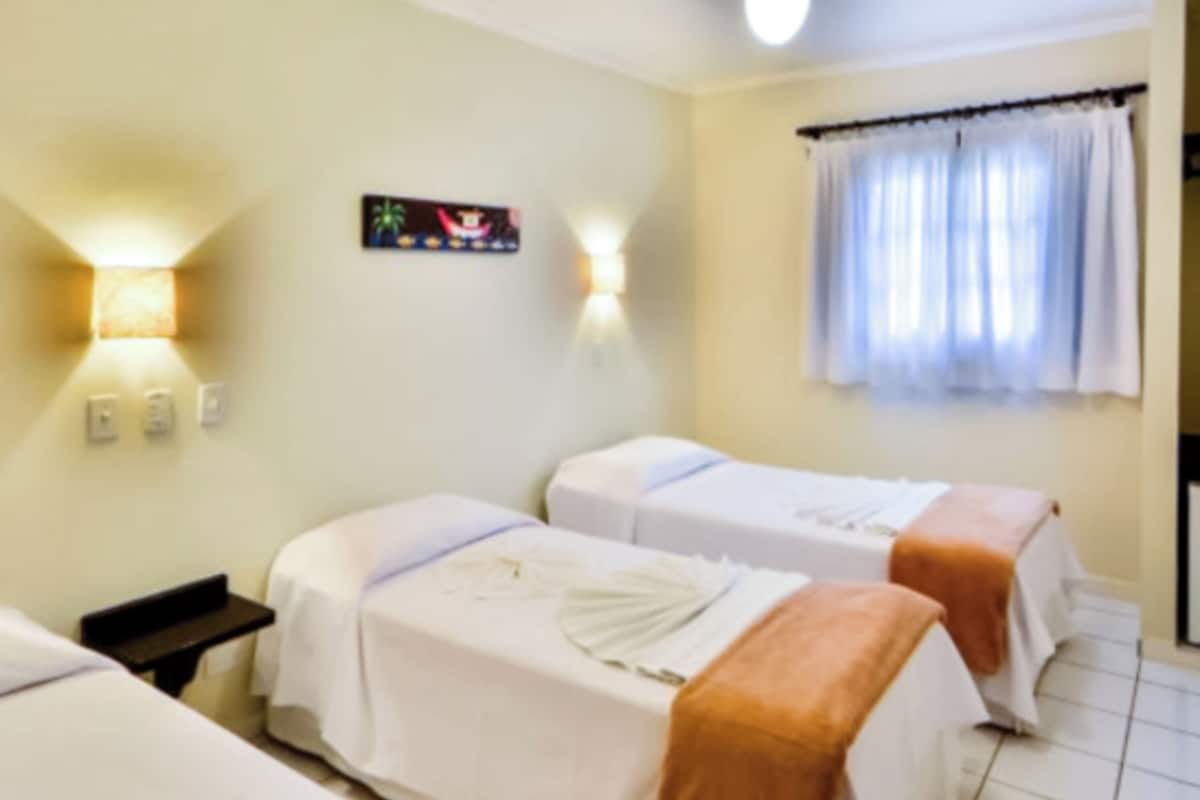 A hotel room with two beds, each covered with white linens and a brown blanket. There are wall-mounted lamps and a window with curtains.