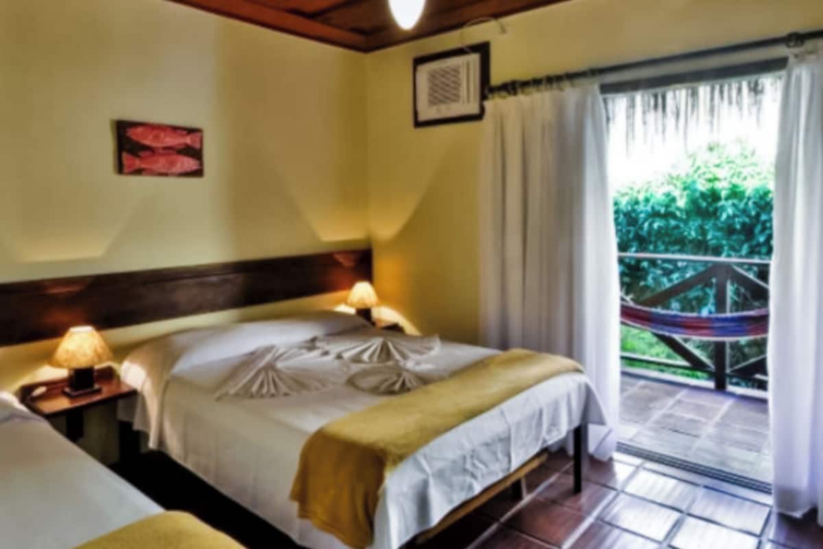 A hotel room with a bed, bedside tables, a window with curtains, and a view of greenery outside.