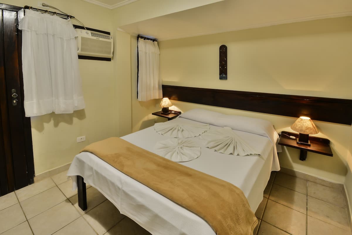 A hotel room with a bed, two bedside tables with lamps, a wall-mounted air conditioner, and a door with a curtain.