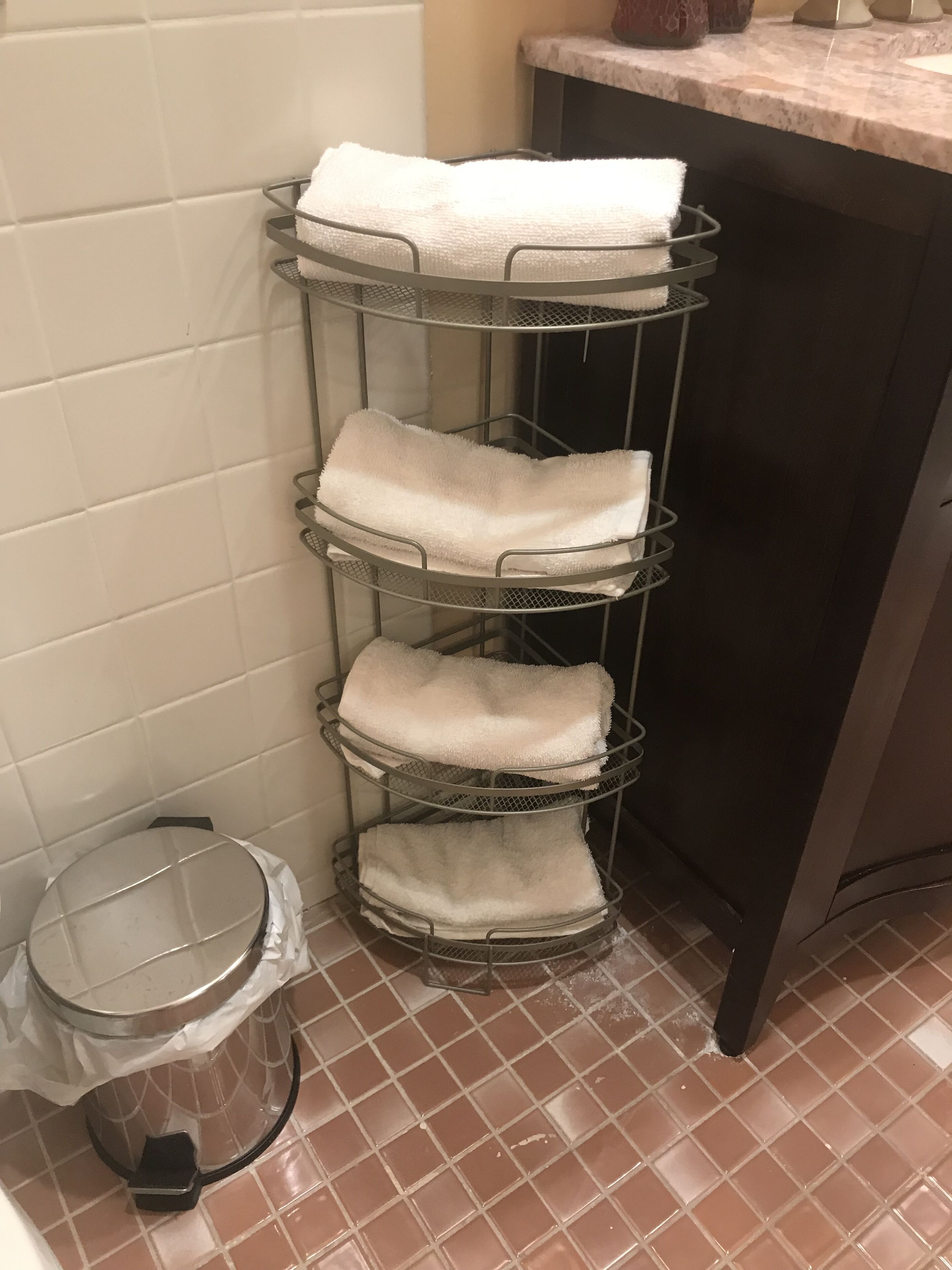 A metal drying rack with white towels against a tiled wall, a trash bin, and a dark countertop.