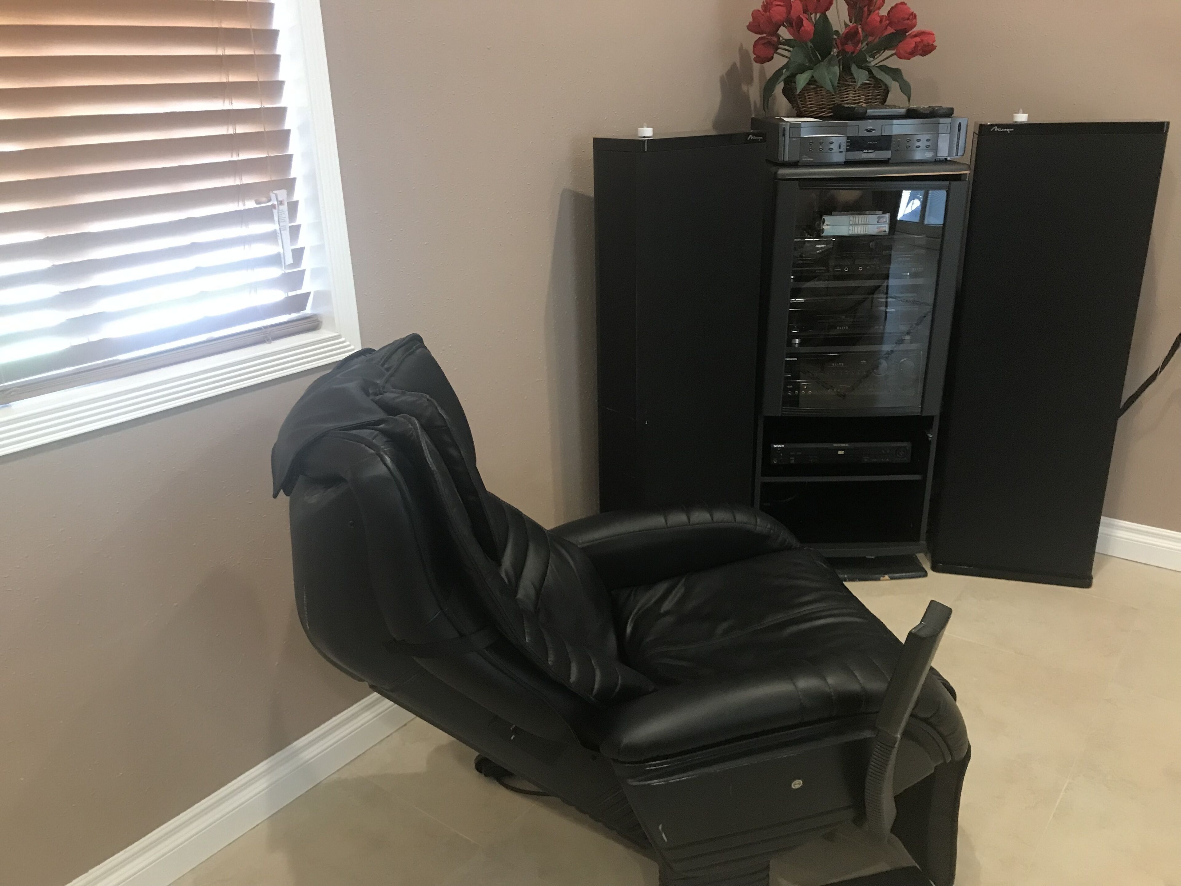 Quarto com uma poltrona de couro preto, um armário preto com dispositivos eletrônicos e uma janela com persianas.