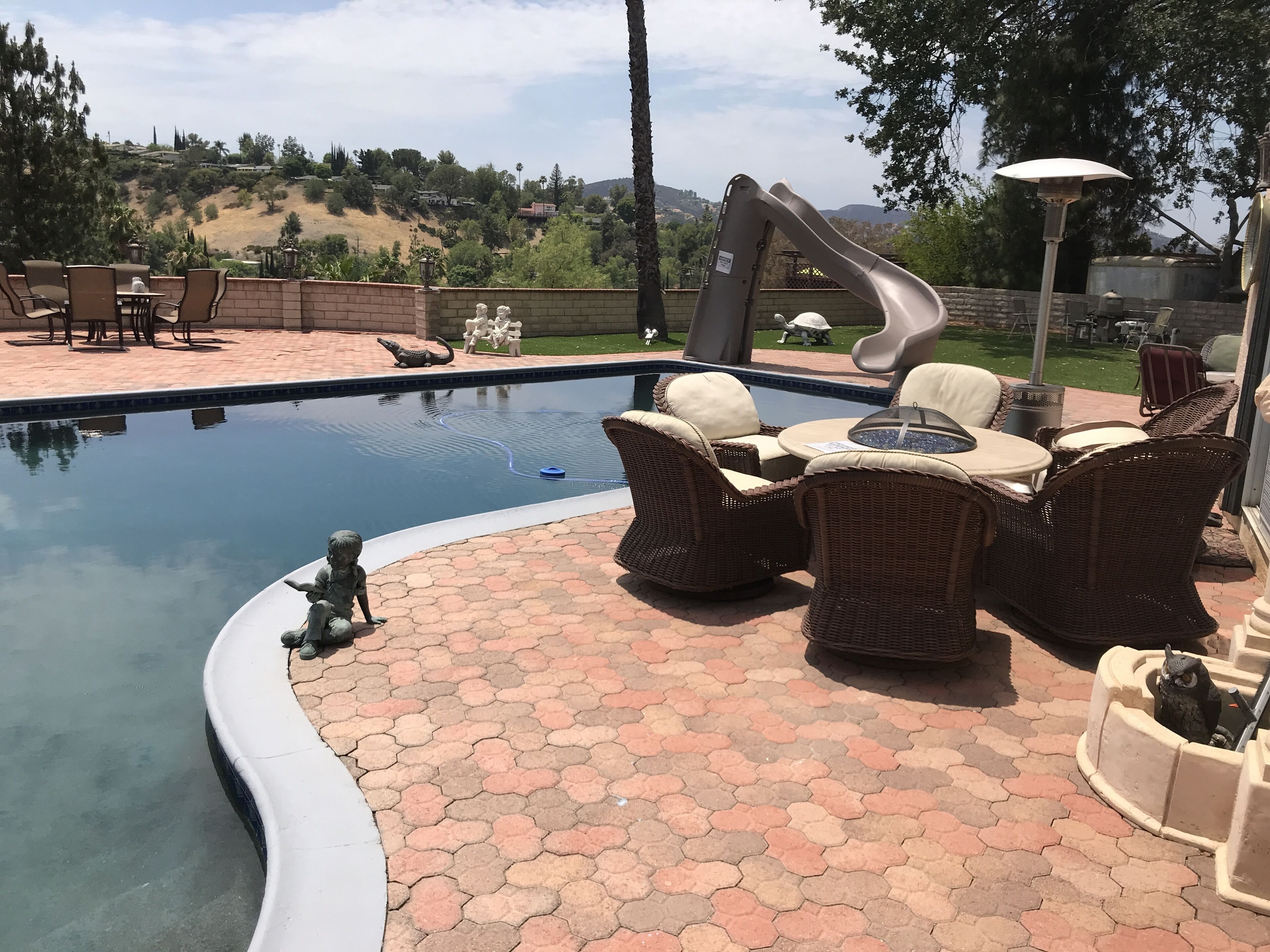 A swimming pool with a curved edge, surrounded by hexagonal tiles. There are wicker chairs and a table with cushions, and a water feature with a blue jet.