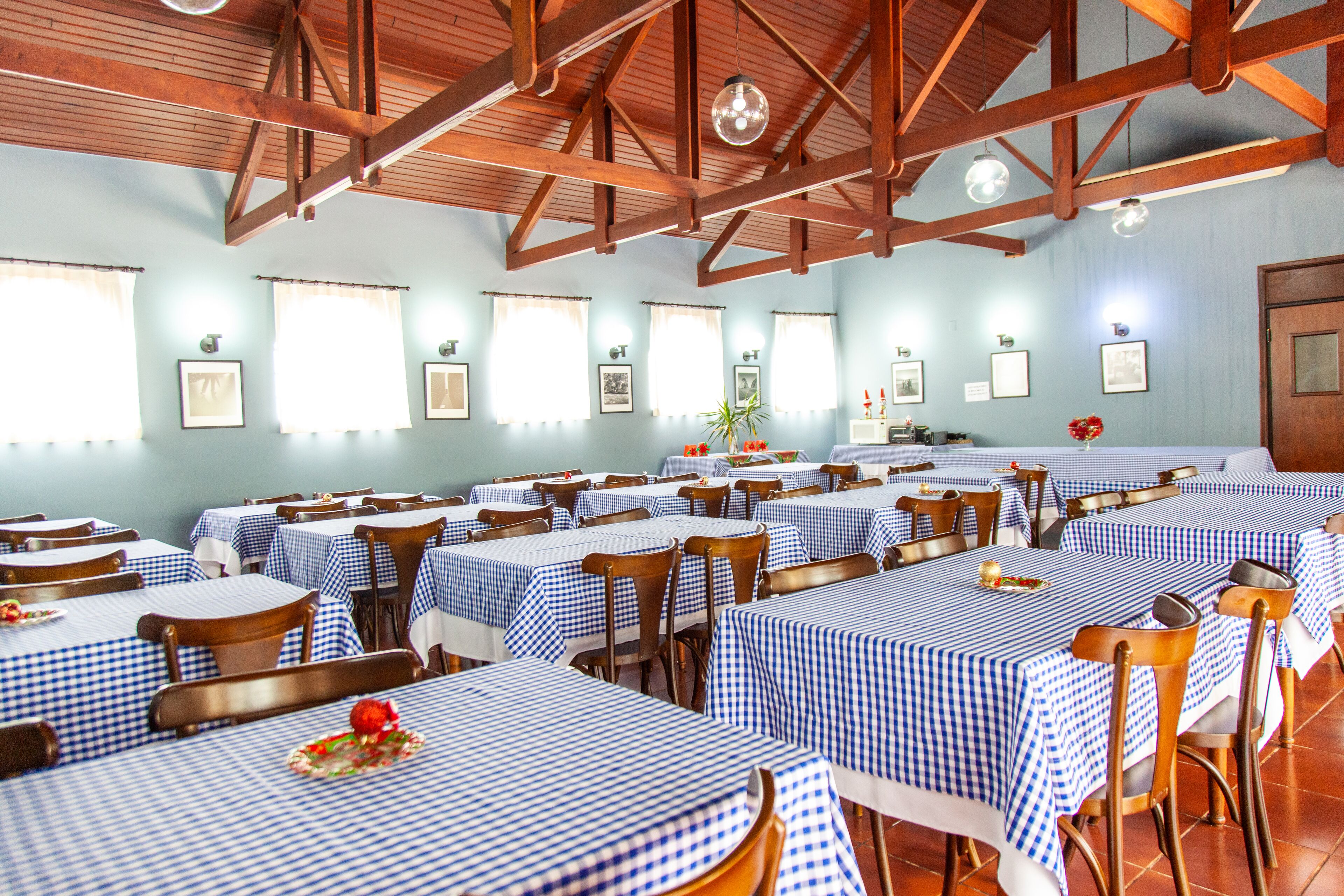 Uma sala de jantar com várias mesas cobertas por toalhas xadrez azuis e brancas, cadeiras de madeira e quadros nas paredes.