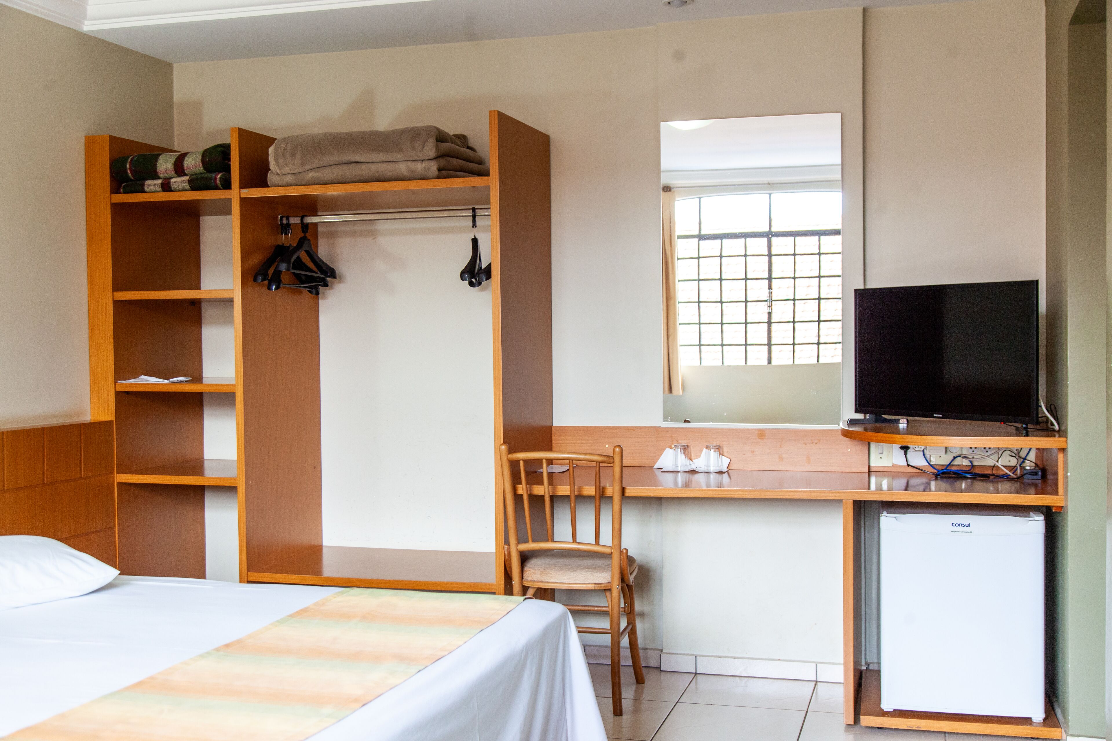 A hotel room with a bed, a wooden wardrobe, a desk with a television, and a small refrigerator.