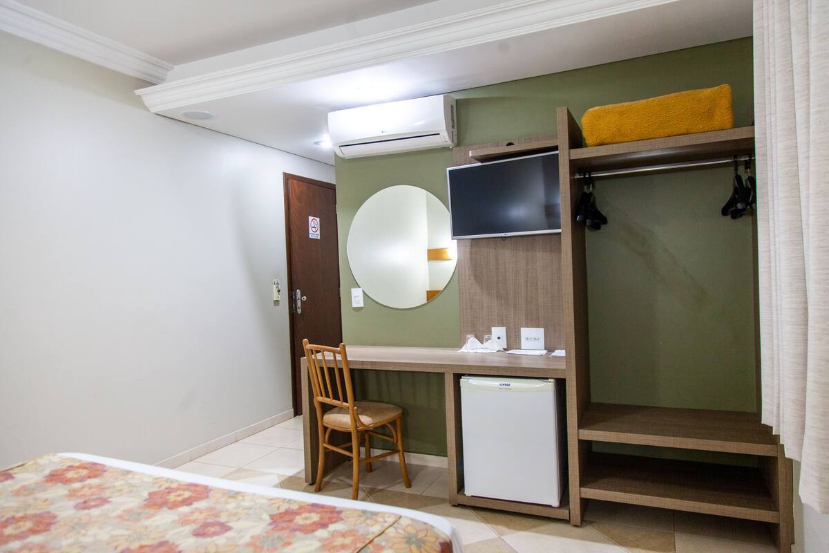 Une chambre d’hôtel comprenant un lit, un bureau avec une chaise, un petit réfrigérateur, un téléviseur fixé au mur, un miroir et une armoire sur laquelle est posée une serviette jaune.