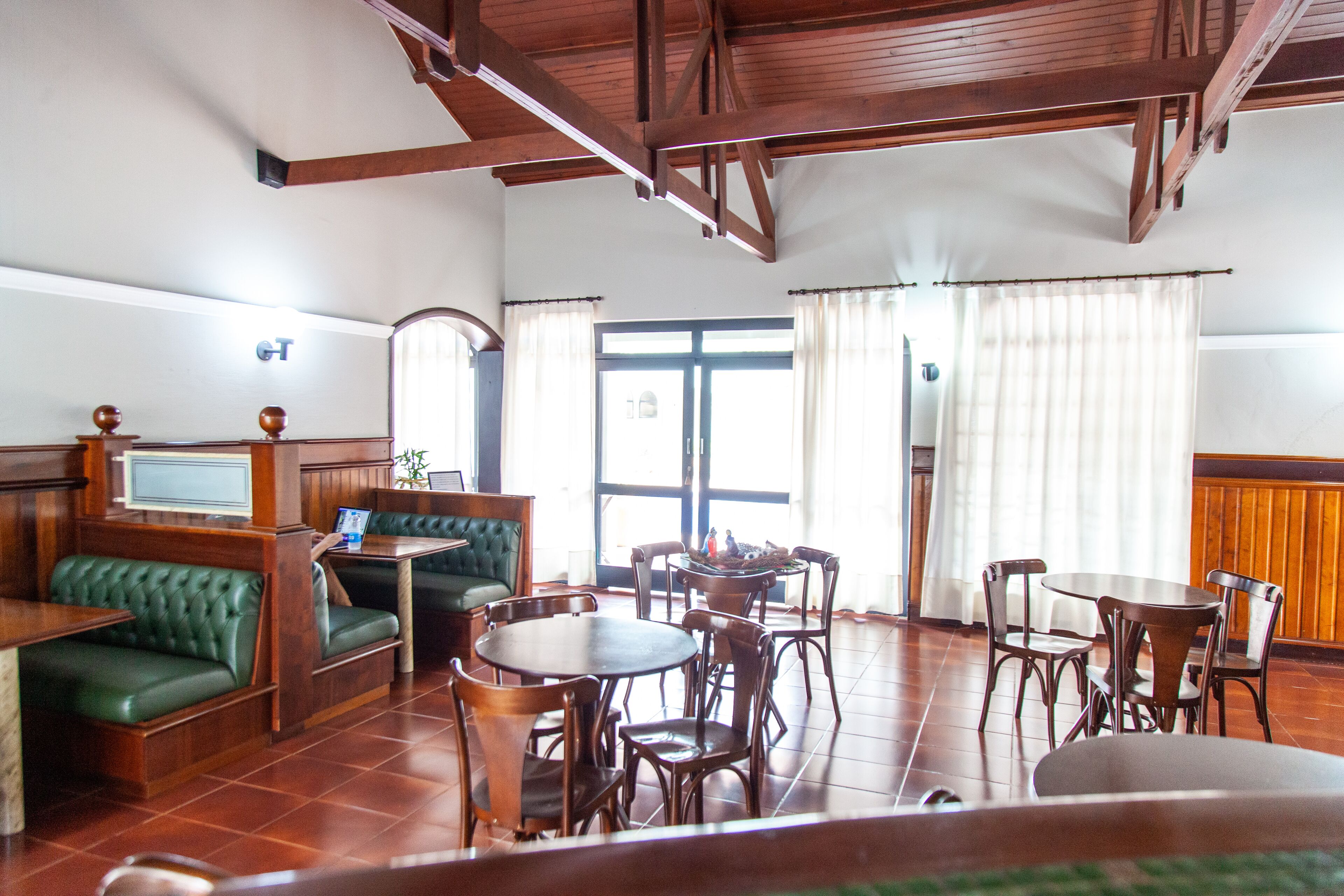 A spacious room with wooden floors, a green cushioned bench, multiple wooden tables and chairs, and a large window with curtains.