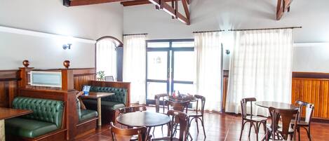 A spacious room with wooden floors, a green cushioned bench, multiple wooden tables and chairs, and a large window with curtains.