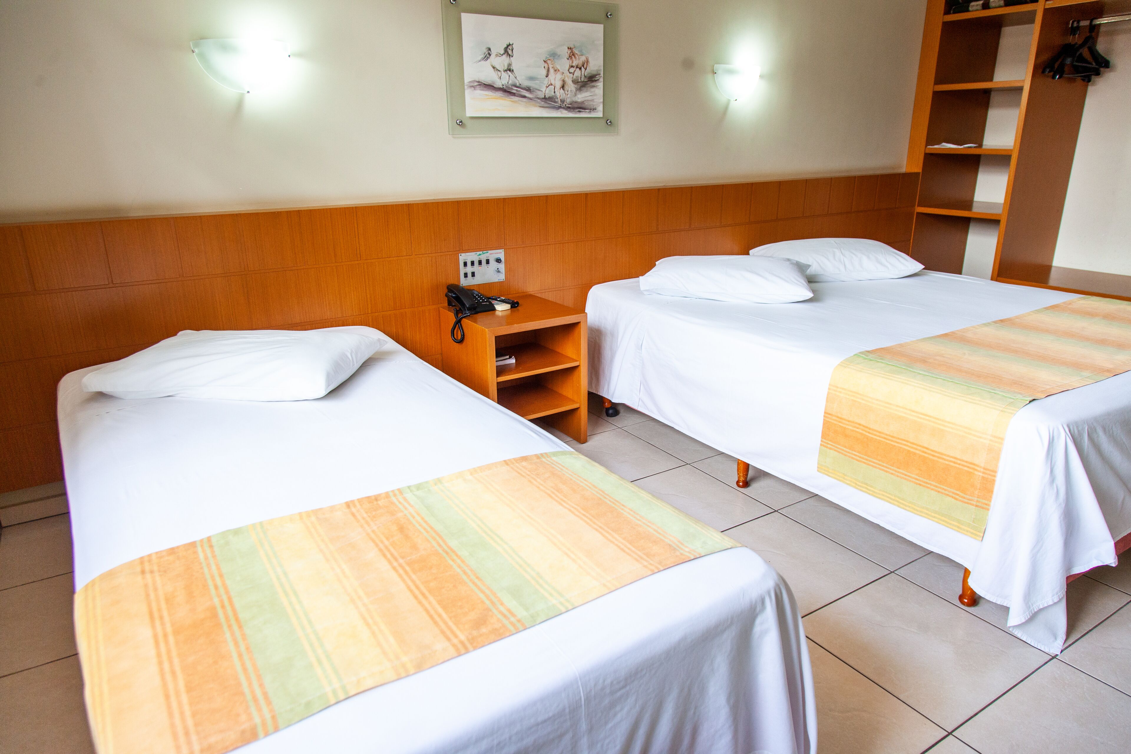 Habitación de hotel con dos camas individuales, una mesita de noche de madera con un teléfono, un cuadro en la pared y un armario con perchas.