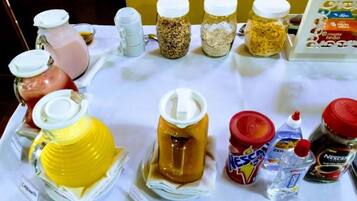 Una mesa con diferentes alimentos y condimentos, entre ellos, frascos de conservas, una jarra de jugo y sobres de café y té.