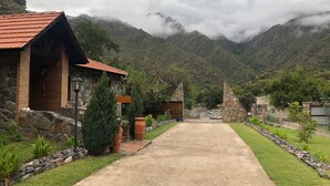 A rustic stone building with a red-tiled roof, a paved pathway leading to a mountain pass, and lush greenery surrounding the area. - Paso del Cóndor (Nogolí)