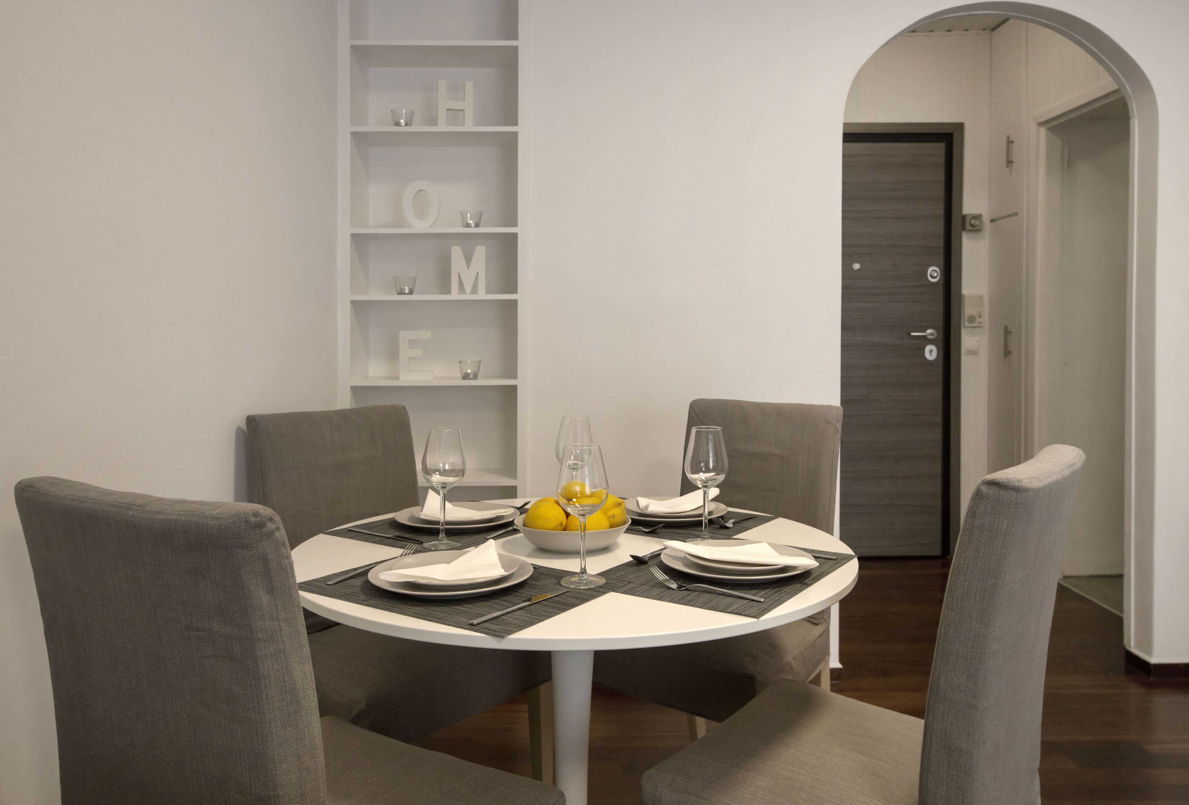 A dining area with a round table set for four, surrounded by grey upholstered chairs. A white shelf with the word "HOME" is visible to the left of the table.