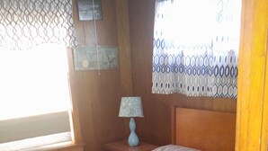 A bedroom with a bed, a nightstand, a lamp, a window with patterned curtains, and a wall with a sign.